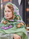 A woman elegantly wearing a thick square scarf made of merino wool, showcasing a floral rose pattern in shades of green, purple, and beige with fringe detailing.