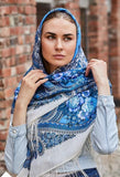Woman wearing a blue floral Slavic headscarf against a brick wall. This square shawl combines a milky white base with light blue floral design for a fresh look.