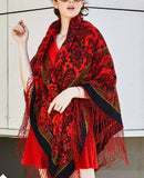 Woman wearing a red and black patterned oversized floral scarf with long fringes in Spanish flamenco style against a light-colored wall. A detailed fashion shot focusing on the front draping of a red wool Slavic scarf on a woman. The intricate traditional design features layers of bright red blossoms, a bold black lace frame, and delicate woven fringe detailing, perfectly accentuating a classy winter fashion accessory.