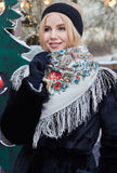 A detailed view of a blonde woman wearing an ornate Slavic-style winter scarf made from 100% merino wool. The heavy, warm ivory fabric is draped closely around her face, highlighting the fine, multicolored paisley scrollwork and the long, flowing white fringe hanging over her shoulder.