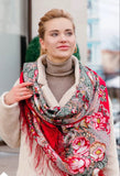 Woman wearing a colorful red floral merino wool scarf with a blurred urban background. A medium close-up of a blonde woman wearing gold hoop earrings, a beige turtleneck sweater, and a plush cream-colored coat. She is wrapped in a large red merino wool shawl decorated with a dense, traditional Slavic pattern featuring large pink roses, small yellow flowers, and sage green leaves, finished with a fine red fringe trim.