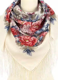Product shot of a large, square merino wool shawl in a traditional Slavic style on a white mannequin. The scarf features a clean ivory base adorned with oversized, vivid pink and coral peonies, stylized light blue flowers, and olive green foliage. The fabric is folded diagonally and draped symmetrically, showing off long, delicate cream fringes along the V-shaped hem.