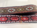 Two rolled widths of patterned woven trim or ribbon with a geometric tribal design in red, black, white, and tan, stacked on a white surface.