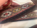 A close-up of a hand holding a roll of decorative woven trim with a geometric diamond pattern in red, gold, black, and white.