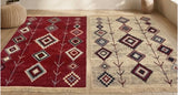 Two rectangular area Armenian rugs with geometric tribal patterns displayed side-by-side on a light wood floor. The left rug is deep red with cream, blue, and green patterns, and the right rug is beige with dark red, blue, and green patterns.