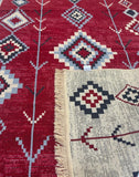 A close-up of a woven rug with a deep red field featuring geometric diamond and arrow-like motifs in cream, navy, and light blue, alongside a cream section with fringe and matching patterned detailing.
