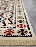 A close-up of a woven rug corner showing cream, red, navy, and beige geometric patterns with fringe along one edge, placed on a light gray wood floor.