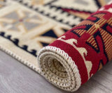 A close-up of a rolled area rug showing its cream-colored twisted edge and a patterned face with red, beige, and navy blue geometric and tribal-style designs, resting on a light gray wood floor.