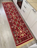 A long red geometric Armenian runner rug with cream, black, and gold patterns and fringe, placed on a light wood floor next to white kitchen cabinets.
