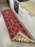 A long rectangular area rug with a red, beige, and black Armenian geometric tribal pattern is shown on a light wood floor in a kitchen setting, positioned under white cabinets and a wooden countertop.