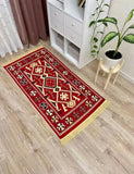 A rectangular red area rug with a Armenian traditional geometric tribal pattern and gold fringe, displayed on a light wood floor in a room setting next to a white bookshelf and a potted plant.