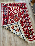 A rectangular woven rug with a bold geometric Armenian pattern, shown partially folded over on a light wood floor. The rug features a dominant deep red field with cream, black, and tan accents in the borders and central medallion, finished with a cream fringe.