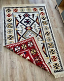 A rectangular woven rug with fringe on two short ends, shown laid on a light wood floor with one corner folded back to reveal the reverse side. The rug has a cream base with bold geometric motifs in black, red, beige, and gray.