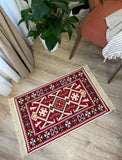 A rectangular red and cream geometric patterned rug with fringe on all sides, laid out on a light wood floor in a styled room corner with a potted plant and an orange armchair visible.