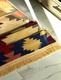 A close-up of a woven rug with a Southwestern geometric pattern in beige, gold, navy blue, red, and orange, featuring a fringed edge, laid out on a white marble floor.