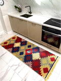 A long rectangular kitchen runner rug with a colorful Armenian geometric diamond pattern and fringe ends, placed on a white marble-tiled kitchen floor in front of beige cabinets and a black oven.