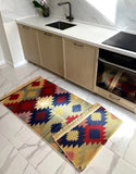 A rectangular area rug with a colorful geometric diamond pattern in red, blue, yellow, and beige, styled on a light marble kitchen floor next to a matching folded rug.