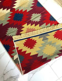 A folded view of a woven area rug with a Armenian geometric pattern in red, mustard yellow, navy blue, and sage green, featuring a fringed edge, shown on a white marble floor.