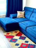 A bright blue sectional sofa with a tufted chaise lounge and a decorative multicolor throw pillow, set in a living room on a patterned geometric Armenian rug in red, yellow, blue and mustard.