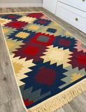 A rectangular woven area rug with a Armenian geometric pattern in navy blue, red, cream, mustard yellow, and sage green, featuring a fringed border, shown on a light wood floor next to a white cabinet.