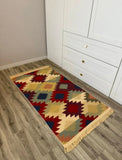 A rectangular area Armenian rug with a colorful geometric kilim pattern, shown on a gray wood-look floor next to a white cabinet.