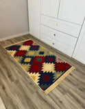 A rectangular area rug with a colorful Armenian geometric kilim pattern and fringe, shown on a gray wood-look floor next to a white cabinet.