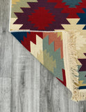 A close-up of a woven rug corner with fringe, showing a geometric Armenian taraz-style pattern in red, navy, beige, and sage green on a gray wood floor.