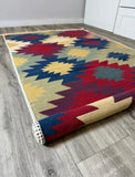 A rolled area rug with a Southwestern geometric pattern in red, navy blue, yellow, and beige, shown on a gray wood-look floor.