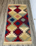 A rectangular woven area Armenian rug with a Southwestern geometric diamond pattern in red, navy, beige, mustard, and sage green, displayed on a gray wood floor.