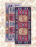 Two rectangular patterned Armenian rugs with fringed edges displayed flat against a floral wallpaper background. The rug on the left has a light blue base with red, navy, and beige geometric motifs.
