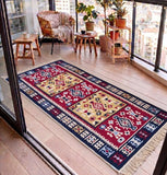 A rectangular area Armenian rug with a detailed geometric and tribal pattern in red, navy blue, beige, and white, styled on a wooden balcony with a wicker chair, plants, and wooden stools.