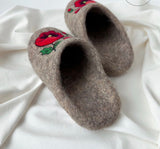 A pair of brown felt slip-on slippers with embroidered red poppies and green leaves, resting on a draped white fabric background.