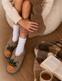Woman wearing woolen slippers with orange berries near the blanket and book.