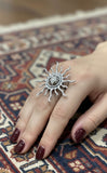Silver sun-shaped ring on a hand with maroon nail polish on a patterned fabric background