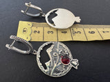 Silver earrings with a red gemstone on a black background with a ruler for scale.