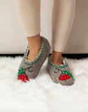 A person wearing beige ribbed leggings stands on a white faux-fur rug, showing a pair of gray felt slip-on slippers decorated with red berries and green leaves.