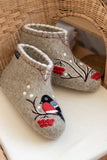 Pair of high shaft bootie-slippers made of merino wool with a design of a bird and berries on a woven chair.