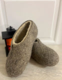 Pair of brown sheep wool clogs slippers on a light wooden floor with a white wall in the background.