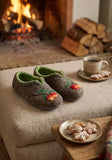 A pair of cozy felt house winter woolen slippers with green lining and embroidered red berries sits on a beige upholstered ottoman in front of a warm fireplace. A cup of coffee and cookies are placed nearby, creating a cozy home atmosphere.
