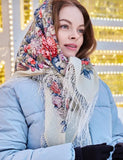 Woman wearing a floral Slavic woolen scarf in white and pink and blue coat with a blurred festive background. Oversized winter head shawl made of premium wool, showcasing intricate folk blue and red flower ornaments and fine ivory fringe borders.