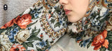 A close-up portrait of a blonde woman looking at the camera, wearing an ivory merino wool shawl styled as a traditional Slavic headscarf. The fabric features an ivory-white base filled with intricate multicolored paisley and floral patterns in red, orange, and blue. The shawl is tied under her chin with long, delicate fringes cascading over her dark winter coat.