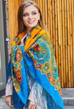 Product lifestyle shot of a woman wearing a multicolored, oversized merino wool palantine. The top portion is styled over her head in bright orange with subtle folk prints, transitioning into a large, warm blue shawl covered in intricate Slavic floral ornaments and finished with an ornate, woven fringe trim.