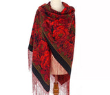 Red floral patterned very large Spanish flamenco scarf in red and black with fringe on a mannequin against a white background. An e-commerce product photograph highlighting a premium red and black Slavic shawl on a model. The blanket-style scarf is wrapped closely around her neck and cascading down her front, showcasing dense, traditional floral arrangements and matching red fringes.