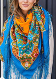 Close-up detail of a woman modeling a luxurious Slavic-style merino wool scarf. The oversized shawl is draped over her shoulders, showcasing a vivid color palette of orange and sky blue adorned with multi-colored folk patterns, complete with long, flowing blue fringe hanging over her light beige coat.