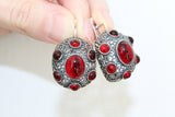 A pair of silver earrings featuring multistone red filigree details, held in a person's hand against a white background.