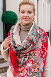Woman wearing a colorful floral merino wool scarf with a red border and large vintage pink flowers with green leaves outdoors. An e-commerce product photograph highlighting a premium red and pink Slavic shawl on a model. The blanket-style scarf is wrapped closely around her neck and cascading down her front, showcasing dense, traditional floral arrangements and matching red fringes.