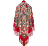 Red floral patterned oversized shoulder shawl made from merino wool with fringes on a white background. A mannequin from the back wearing a warm, blanket-style winter shawl made of premium merino wool. The garment features a rich, traditional Slavic folk pattern filled with pink roses and red fringed borders and a silky fringe on a trim.