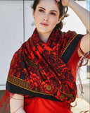 Woman wearing a red and black patterned large wrapping woolen scarf with red roses and fringe. A woman poses while wearing a warm, blanket-style winter shawl made of premium merino wool. The garment features a rich, traditional Slavic folk pattern filled with red roses and a black floral motif on a vibrant red background, draped loosely over a matching red outfit.