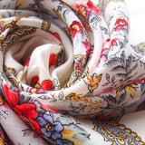 Roll of white floral patterned merino wool shawl with red, yellow, and blue colors. A detailed close-up shot of a luxurious Slavic-style merino wool palantine. The oversized scarf displays a rich botanical border design filled with blooming red roses and complex yellow paisley scrollwork, draped to reveal the folds of the thick fabric.