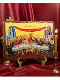An Orthodox Christian icon of the Last Supper on a gold leaf background, displayed on a wooden stand with a red satin backdrop and a lit candle in a brass holder.